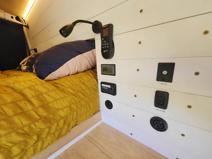 RV rental interior with a cozy bed, white shiplap walls, and a control panel featuring power monitor, inverter, and switches.. Ford Transit 2022