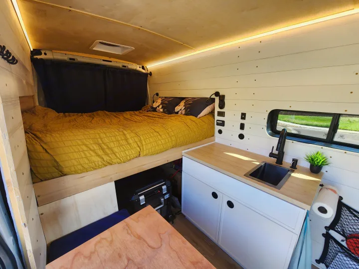 An RV rental's clever interior design shows a comfortable bed, functional kitchenette with sink, wood accents, and bright lighting.. Ford Transit 2022