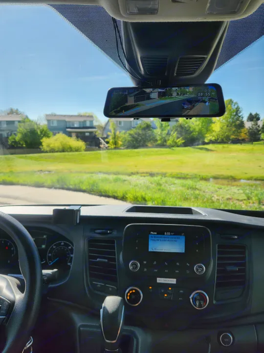 Driver's perspective inside an RV rental. View of dashboard, digital rearview mirror (showing street), and infotainment screen on a sunny day.. Ford Transit 2022