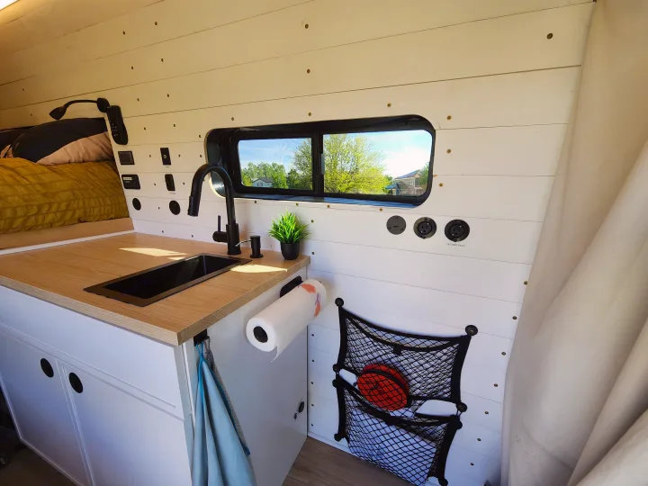 RV rental van's custom kitchenette: black sink, wood counter, wall controls, and practical storage net.. Ford Transit 2022