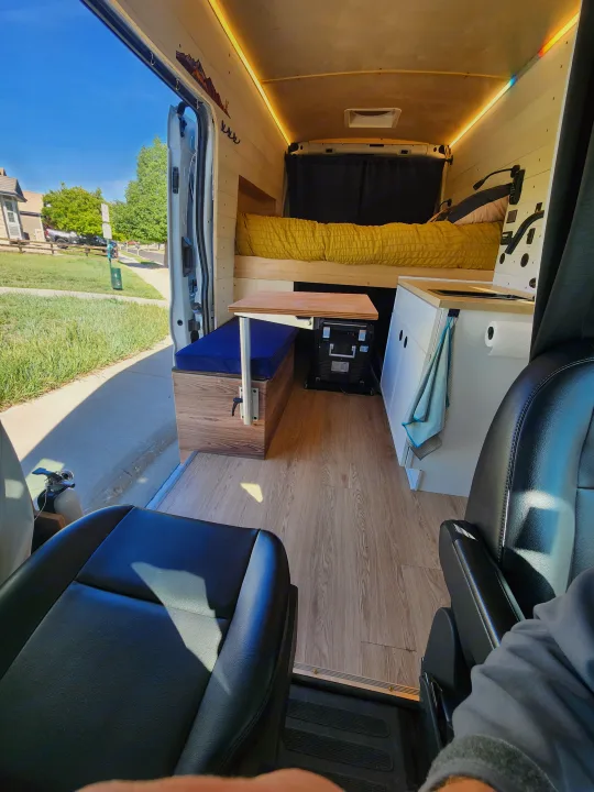 Step into this cozy RV rental camper van: bed, dinette, and kitchen. Wood-paneled interior, with an open door looking outside.. Ford Transit 2022