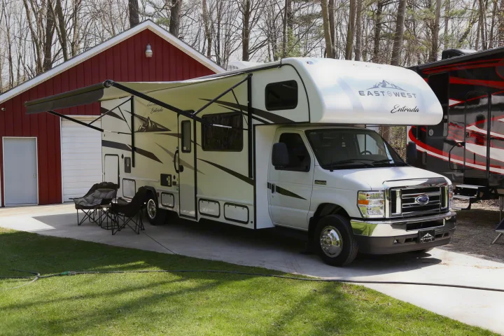 White Class C RV rental with awning extended, parked near a red building and camping chairs, ready for outdoor adventure.. East to West Entrada 2023
