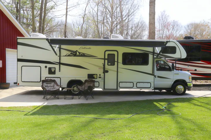 This white Class C motorhome rv rental is parked with its awning extended, chairs set up, and a red building on the left.. East to West Entrada 2023