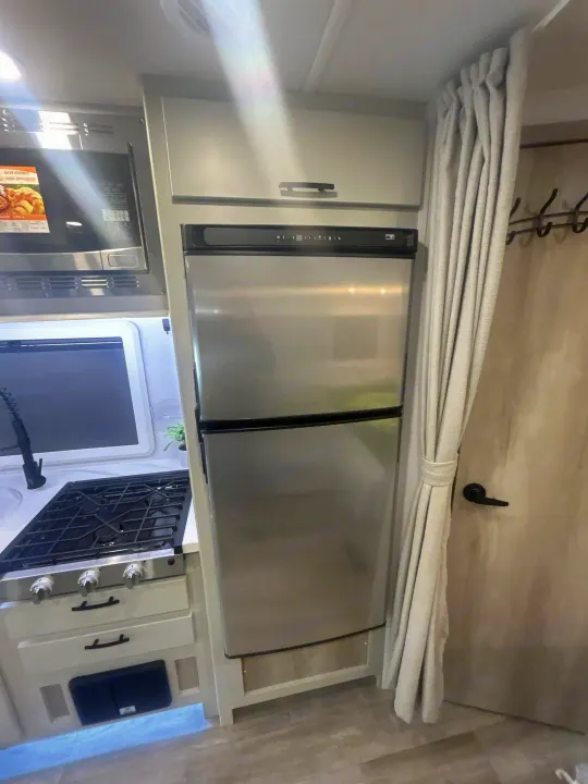 This RV rental's compact kitchen boasts a stainless steel fridge, microwave, and stovetop. Light-colored curtains on the right.. Isata 3 Series 2026