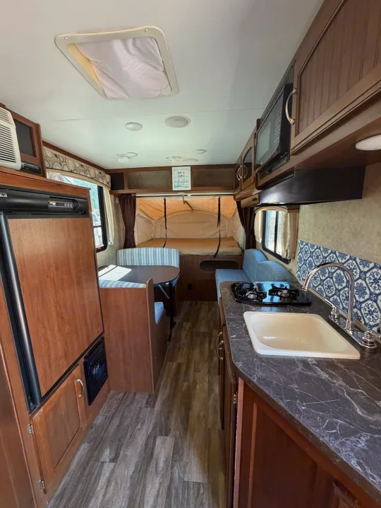 Inside an rv rental: kitchen with sink & stove, dinette, and a pop-out bed. Features wood cabinets & blue accents.. Jayco Jay Feather 2017