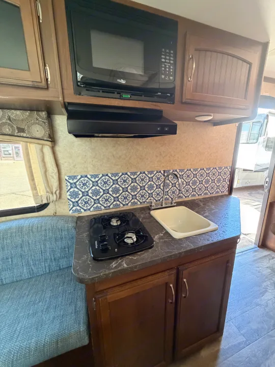 RV rental kitchen: brown cabinets, dark counter, microwave, two-burner stove, sink, and striking blue & white backsplash.. Jayco Jay Feather 2017