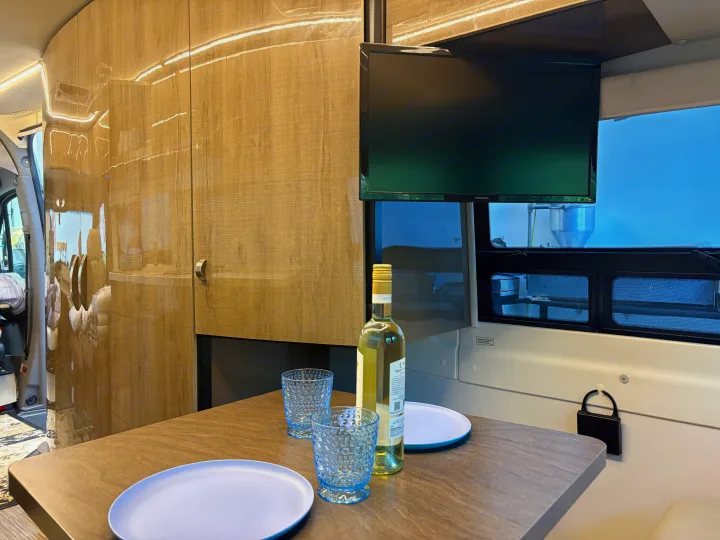 Inside an rv rental: a dining table with wine, glasses, and plates. A TV is mounted above. Light wood walls create a cozy atmosphere.. Winnebago Era 2017