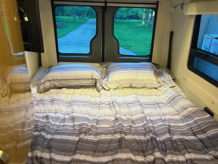 Cozy RV rental bed with striped bedding and a view of a park-like setting from the rear windows.. Winnebago Era 2017