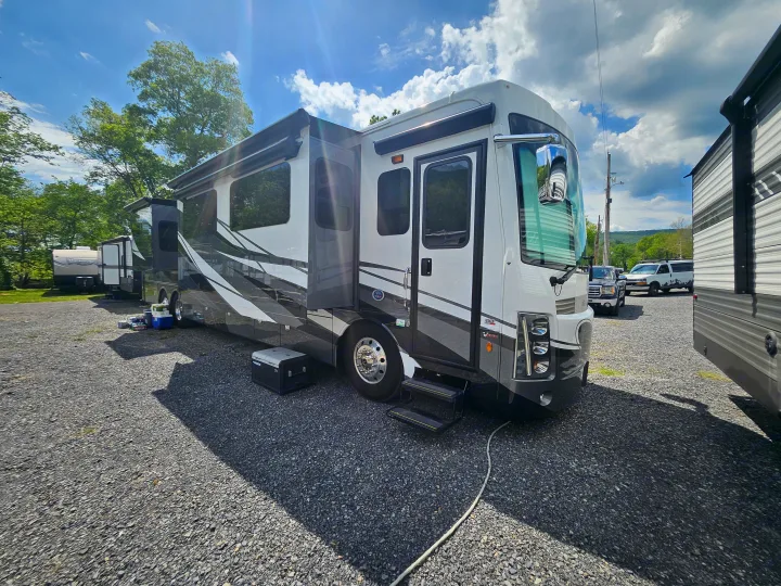 Our RV rental, a large motorhome with extended slides, parked on a gravel campsite under a sunny sky.. Forest River Berkshire XLT 2024
