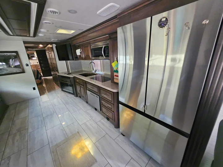 Modern rv rental kitchen featuring stainless steel fridge, microwave, sink, dark cabinets, and light tiled flooring. A cozy, functional space.. Forest River Berkshire XLT 2024