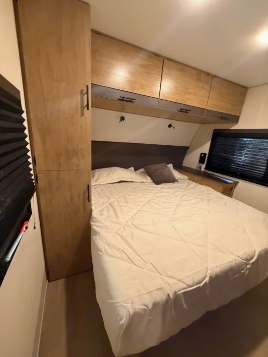 Neatly made bed with light bedding in a modern RV rental bedroom, featuring wooden cabinets, a wardrobe, and black blinds.. Forest River Campsite Reserve 2026