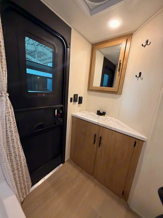 RV rental bathroom: triangular vanity with white sink, wood-grain cabinets, mirrored medicine cabinet, black door, and wall hooks.. Forest River Campsite Reserve 2026