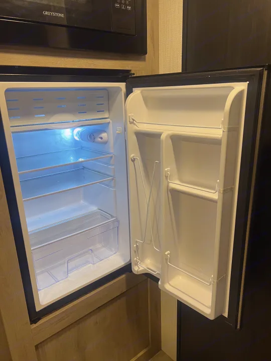 Open, empty mini-fridge with shelves and door storage in an rv rental kitchenette, with a GREYSTONE microwave overhead.. Heartland Prowler Lynx 17BHX 2025