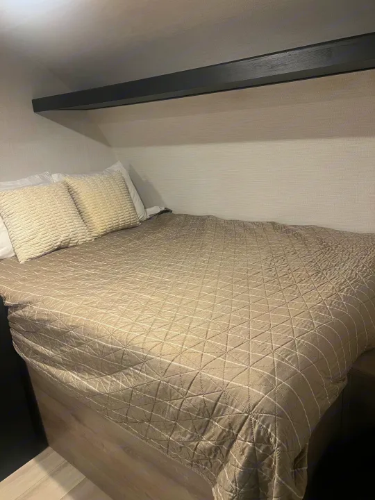 Neatly made bed in an RV rental, featuring light textured pillows, a grey quilted comforter, and a dark wooden overhead shelf.. Heartland Prowler Lynx 17BHX 2025