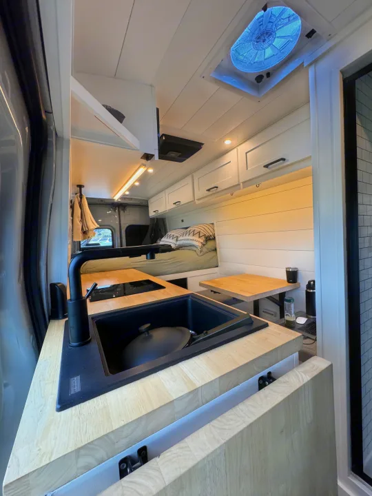 Modern RV rental interior featuring a sleek kitchen with black sink, induction cooktop, light wood counters, and a cozy bed.. ActiVan Ford Transit 350 AWD 2023