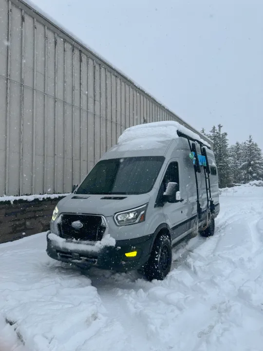 This RV rental is ready for winter adventure! A grey camper van covered in snow, with falling snowflakes, parked by a building.. ActiVan Ford Transit 350 AWD 2023