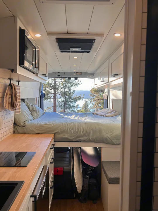Cozy rv rental interior: van bed overlooks a scenic lake & pine trees, with a compact kitchen in foreground.. ActiVan Ford Transit 350 AWD 2023