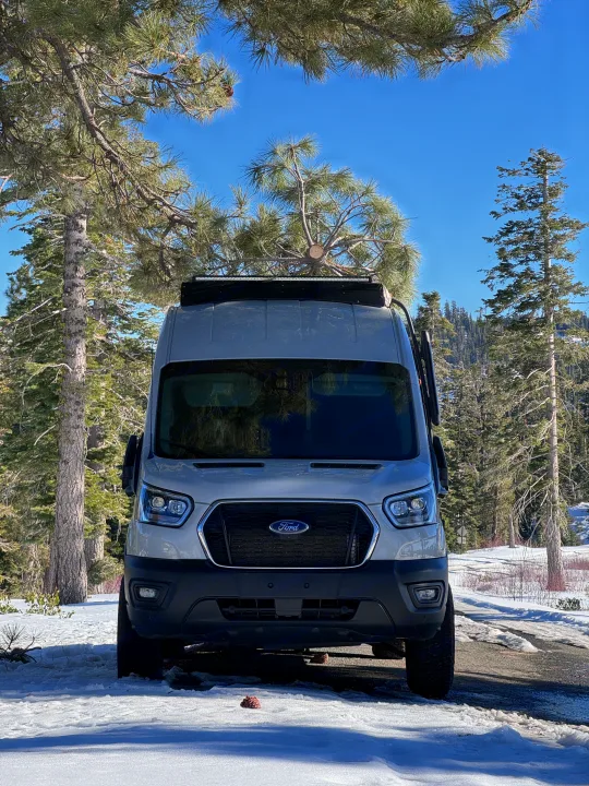 RV rental ready Ford Transit camper van parked in a sunny, snowy forest. Tall pine trees frame the scene under a blue sky.. ActiVan Ford Transit 350 AWD 2023