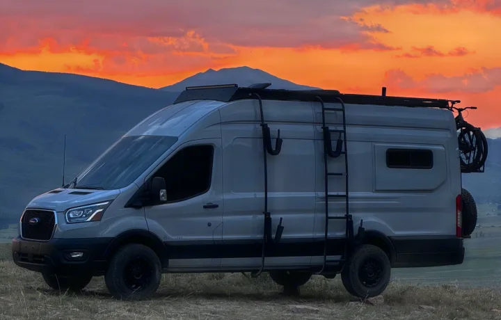 A gray Ford Transit camper van RV rental, equipped for adventure, is parked on a hill with mountains under a vibrant sunset.. ActiVan Ford Transit 350 AWD 2023