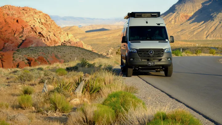 A modern RV rental adventure van on a scenic desert road, surrounded by red rock mountains and arid vegetation.. Other Other 2026