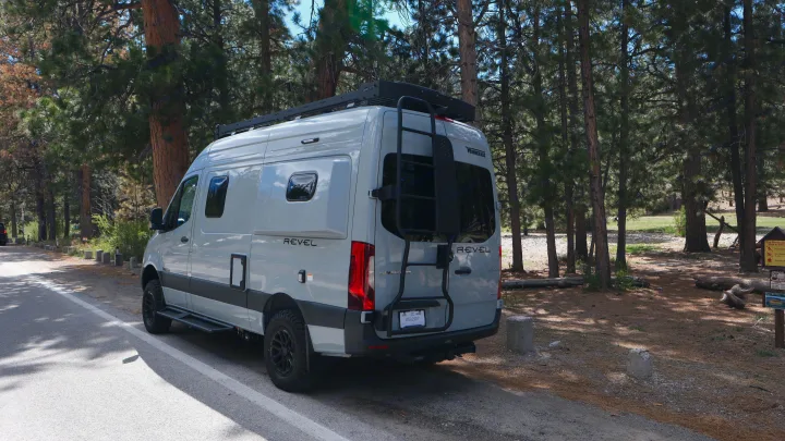 RV rental: A light gray Winnebago Revel camper van, with roof rack and rear ladder, parked by a pine forest.. Other Other 2026