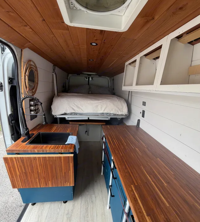 Modern rv rental van interior featuring a comfortable bed, wood-plank ceiling, white walls, and a well-equipped kitchenette.. Mercedes Sprinter 2010