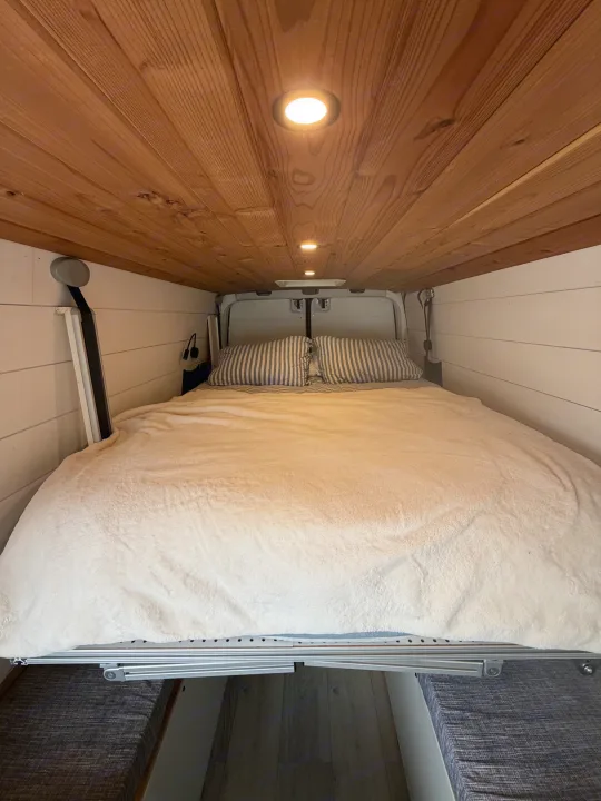 Cozy RV rental interior shows a large bed with striped pillows, a wooden ceiling with lights, and white paneled walls.. Mercedes Sprinter 2010