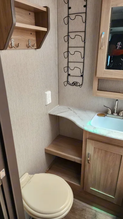 RV rental bathroom featuring a toilet, sink vanity with mirror, wall shelves, and a tall metal storage rack. A compact, functional space.. Coachmen 292bhs 2021