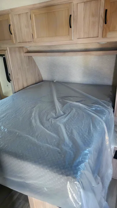 New rv rental bedroom with a bed covered in protective plastic, overhead light wood cabinets, and shelf.. Coachmen 292bhs 2021