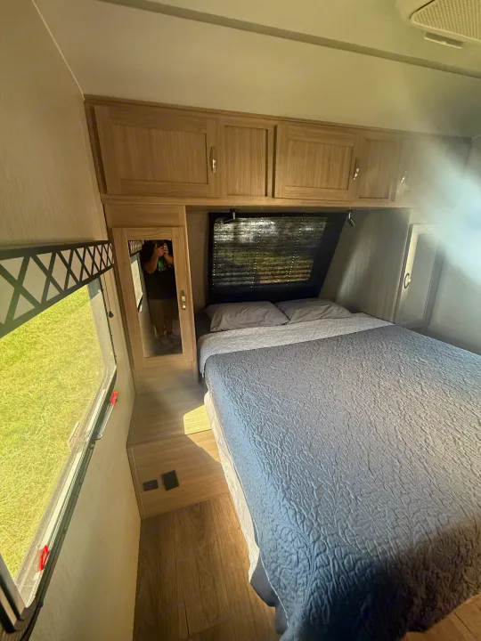 RV rental bedroom: gray bed, wood cabinets, window showing grass. Photographer visible in mirror.. Coachmen Other 2021