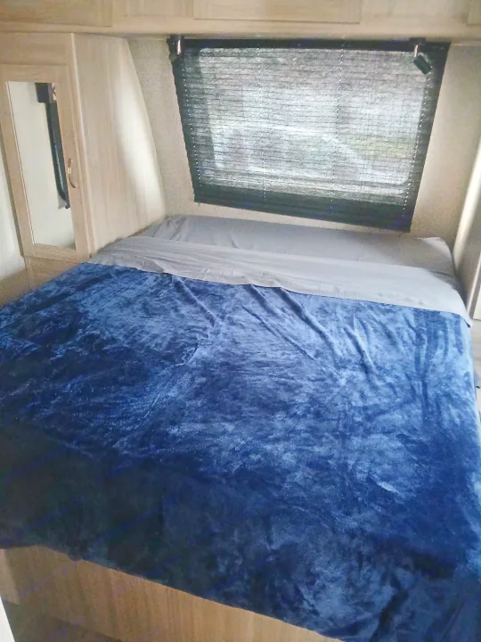 Inside an RV rental: a comfy bed with a plush blue blanket, grey sheets, and window blinds.. Coachmen Other 2021