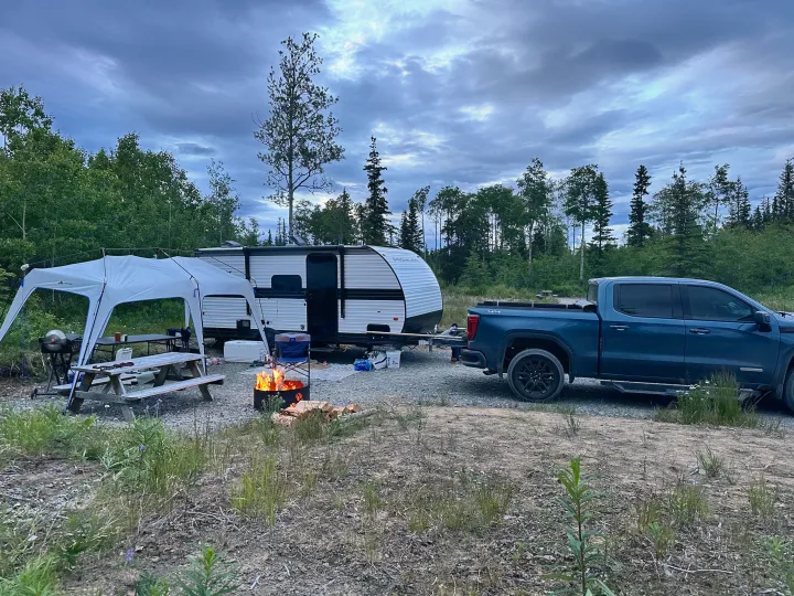 A blue truck pulls an RV rental to a campsite with a lit campfire, picnic table under a canopy, and surrounding trees.. Heartland RVs Prowler Lynx 2024