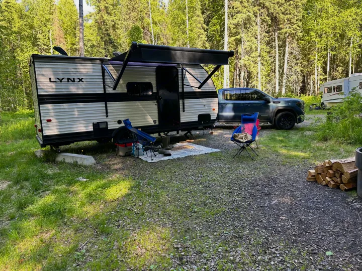 RV rental: Lynx travel trailer & truck parked at a serene forest campsite with chairs, firewood, and another RV.. Heartland RVs Prowler Lynx 2024