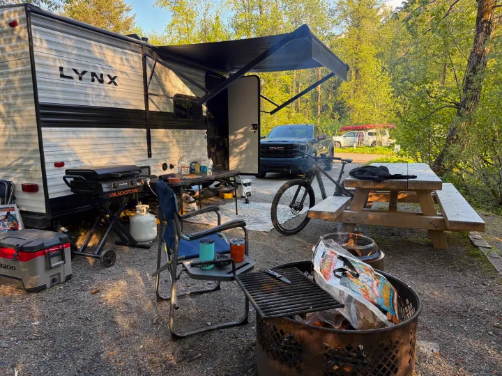 RV rental camping spot with Lynx trailer, GMC truck, Blackstone grill, fire pit, bike, and picnic table in a woodsy setting.. Heartland RVs Prowler Lynx 2024