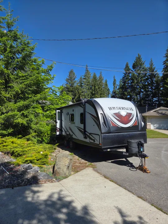 A white and gray Wilderness travel trailer, ideal for an rv rental, parked on asphalt with green trees under a blue sky.. Heartland RVs Wilderness 2018