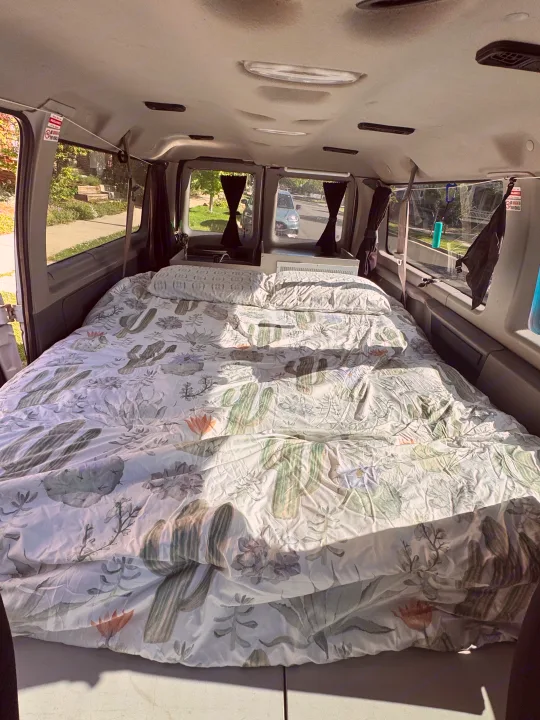 Inside an RV rental: a cozy camper van bed with a unique cactus print comforter, offering a comfortable space for road trips.. Ford Econoline 150 2012
