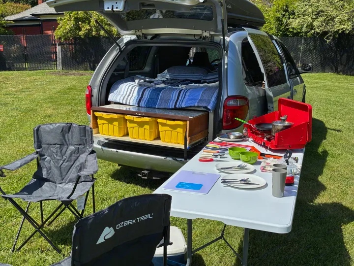 RV rental minivan converted for camping: bed, storage, and an outdoor kitchen with stove, plates, and chairs set up on green grass.. Dodge Grand Caravan 2005