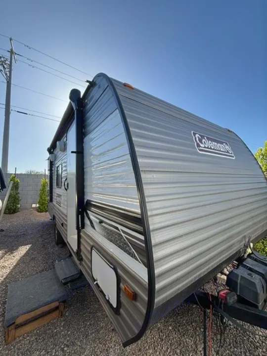Discover this gray Coleman travel trailer RV rental parked on gravel under a bright blue sky, featuring an awning and steps.. Dutchmen Coleman Lantern LT 2024