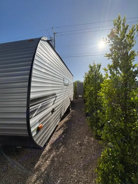 Experience a sunny adventure with this RV rental! A sleek travel trailer sits on gravel, bordered by green hedges.. Dutchmen Coleman Lantern LT 2024