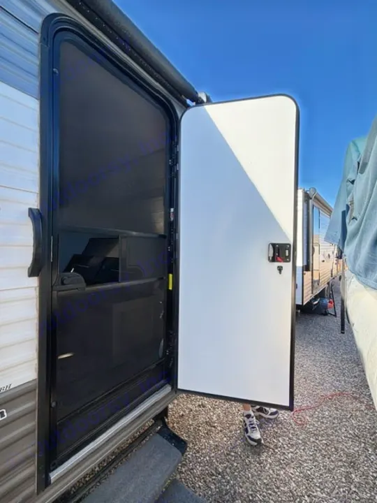 Open RV rental door & screen door reveal interior view. A person's feet are on the gravel. Ready for travel!. Dutchmen Coleman Lantern LT 2024