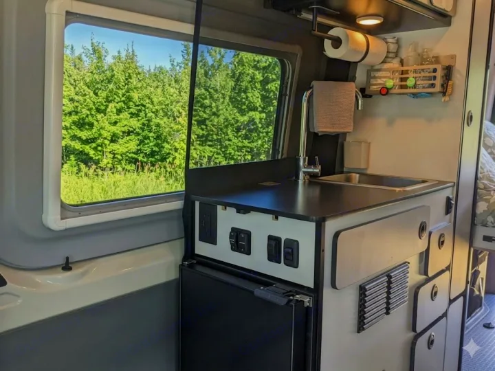 Compact RV rental kitchen with sink, fridge, and forest view for your van life adventure.. Winnebago Revel 2023