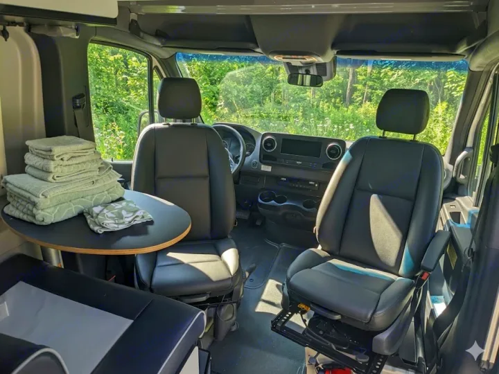 RV rental interior: driver/passenger seats, round table with towels, dashboard, & forest view. Ready for your adventure!. Winnebago Revel 2023