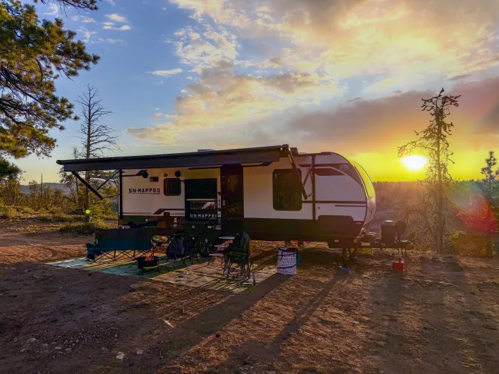 Golden hour RV rental camp with a travel trailer, campfire, and seating nestled among trees.. Forest River Unmapped 2026