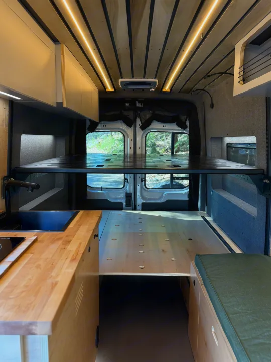 Stylish RV rental interior: high bed platform, sleek kitchen, and cozy bench. Enjoy custom van life adventures.. Ford Transit 2025