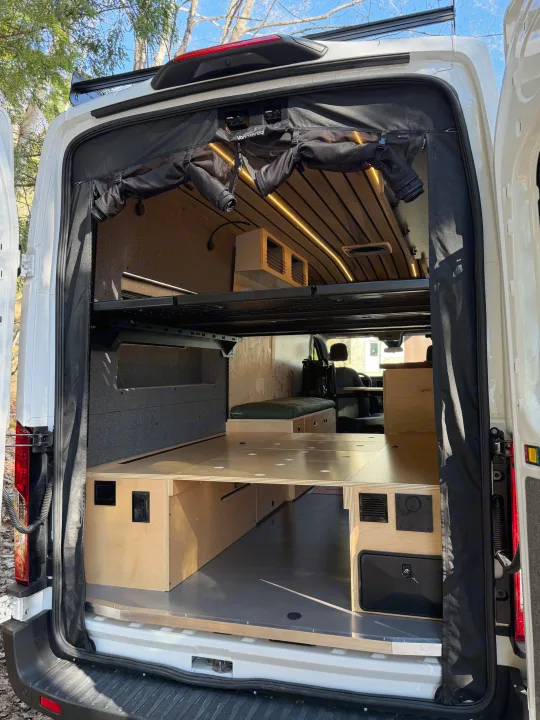 Inside a custom camper van conversion, ideal for an rv rental. Shows a convertible bed, storage, and ceiling lights from the rear.. Ford Transit 2025