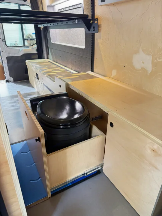 RV rental: Pull-out composting toilet in custom plywood cabinetry of a camper van interior, designed for mobile living.. Ford Transit 2025
