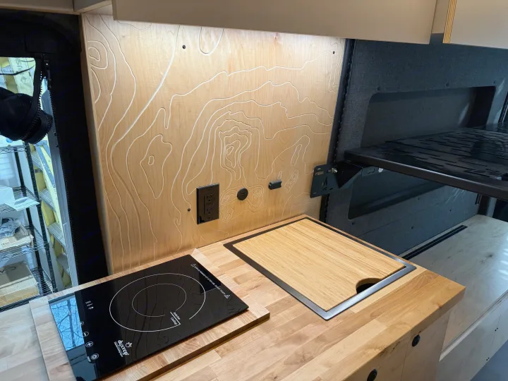 Camper van kitchen with wood counters, induction cooktop, sink, and etched topographic backsplash. Ideal for RV rental.. Ford Transit 2025