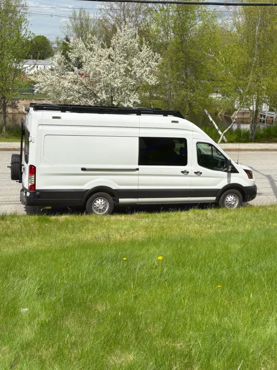 White Ford Transit campervan (RV rental) parked by lush green grass. Blooming trees, houses, and water are in the scenic background.. Ford Transit 2025