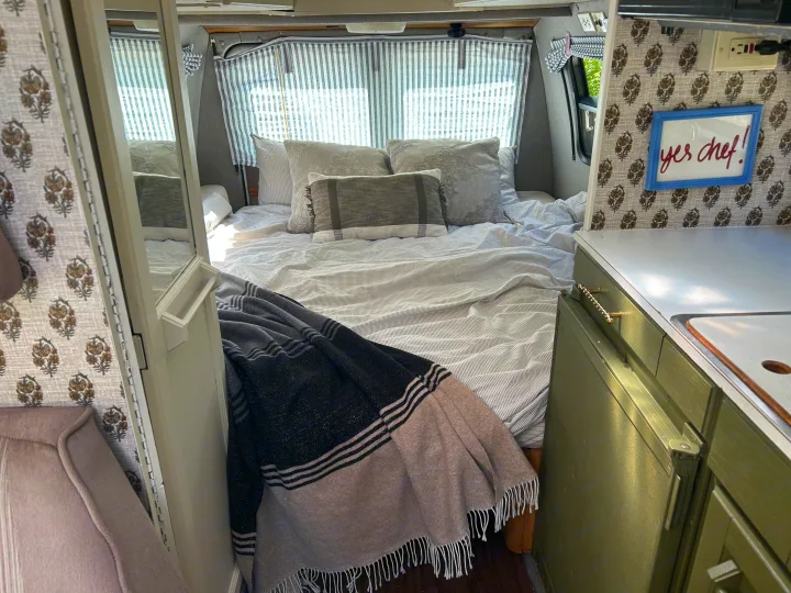 Cozy RV rental interior showcasing a comfortable bed with striped bedding, curtains, and a compact kitchen with a "yes chef!" sign.. Roadtrek 190 Popular 1995