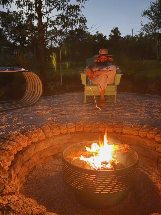An individual in a hat relaxes by a glowing fire pit, perfect after an RV rental adventure. Outdoor evening scene with trees.. Airstream Flying Cloud 2019